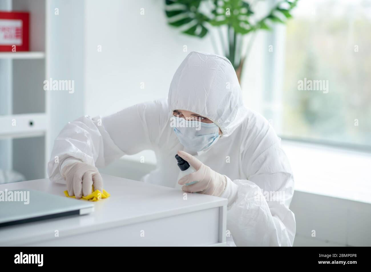 Medical worker in protective clothing and medical mask sprays disinfectant on table Stock Photo