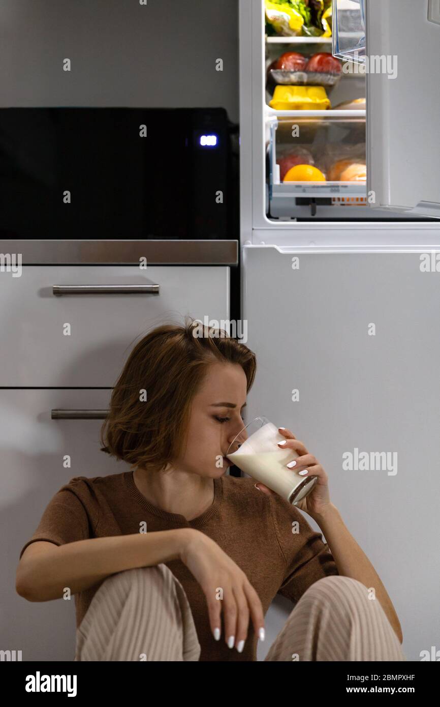 Young woman drinking milk near open refrigerator at midnight while sitting on floor in kitchen. Night eating syndrome Stock Photo