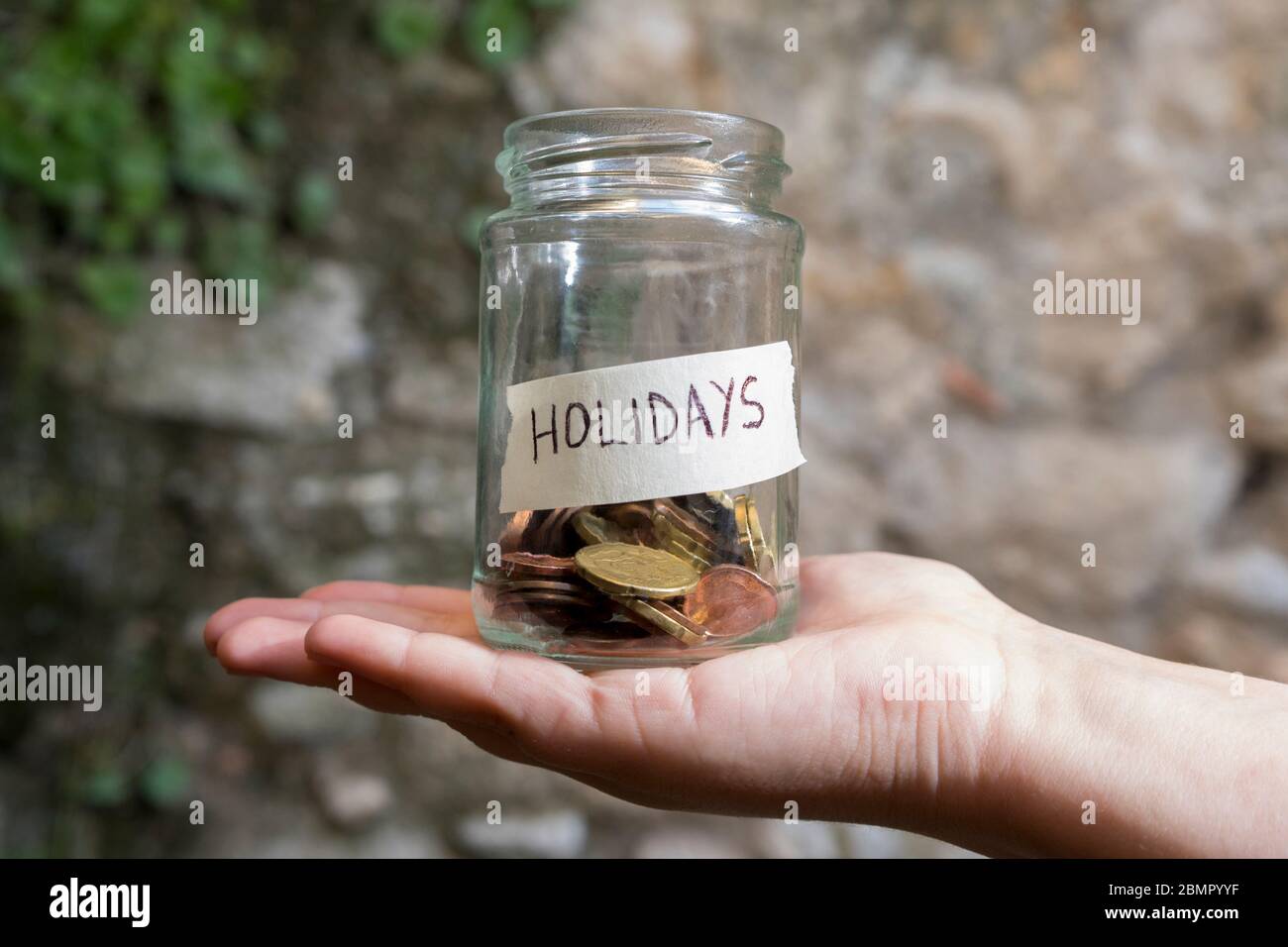 A glass jar with coins inside and a holidays tag on a hand, on a plants and stone wall background. Un bote de cristal con monedas dentro, tiene una et Stock Photo