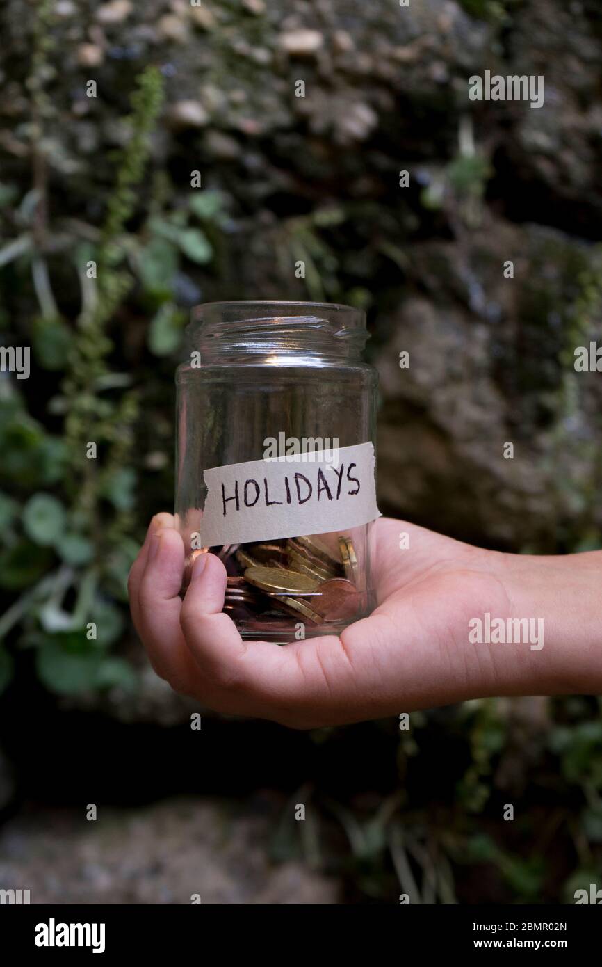 A glass jar with coins inside and a holidays tag on a hand, on a plants and stone wall background. Un bote de cristal con monedas dentro, tiene una et Stock Photo