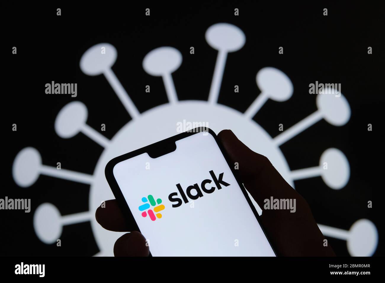 Slack logo on a smartphone silhouette hold in hand. Coronavirus image on the blurred background. Real photo, not a montage. Stock Photo