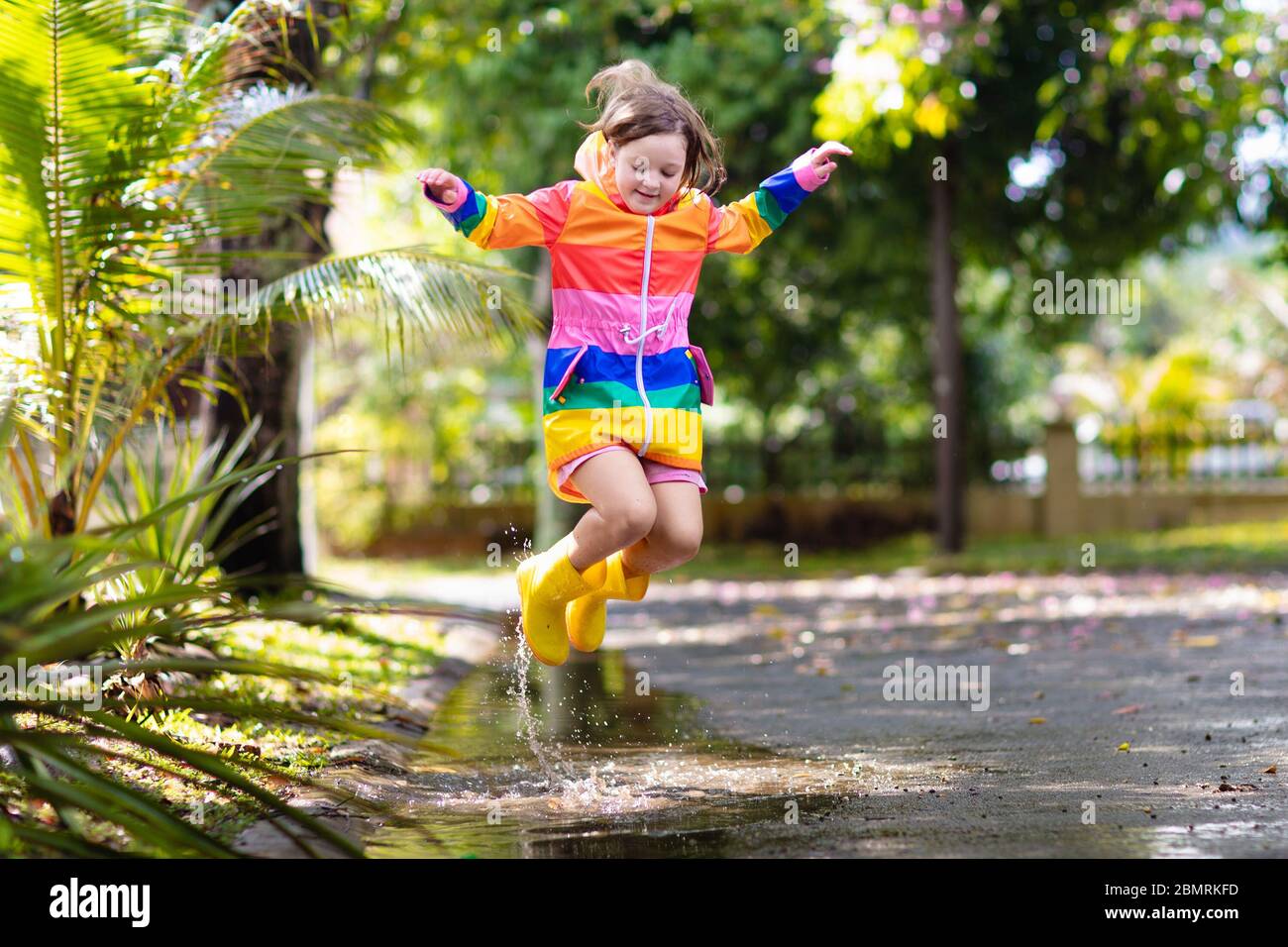 Kid playing in the rain in autumn park. Child jumping in muddy puddle on rainy fall day. Little girl in rain boots and rainbow jacket outdoors in heav Stock Photo