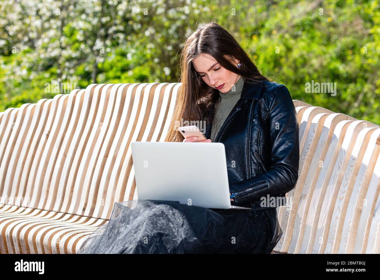 young elegant woman sitting on bench in park and working on laptop on a sunny and windy day, the concept of working outside the office Stock Photo