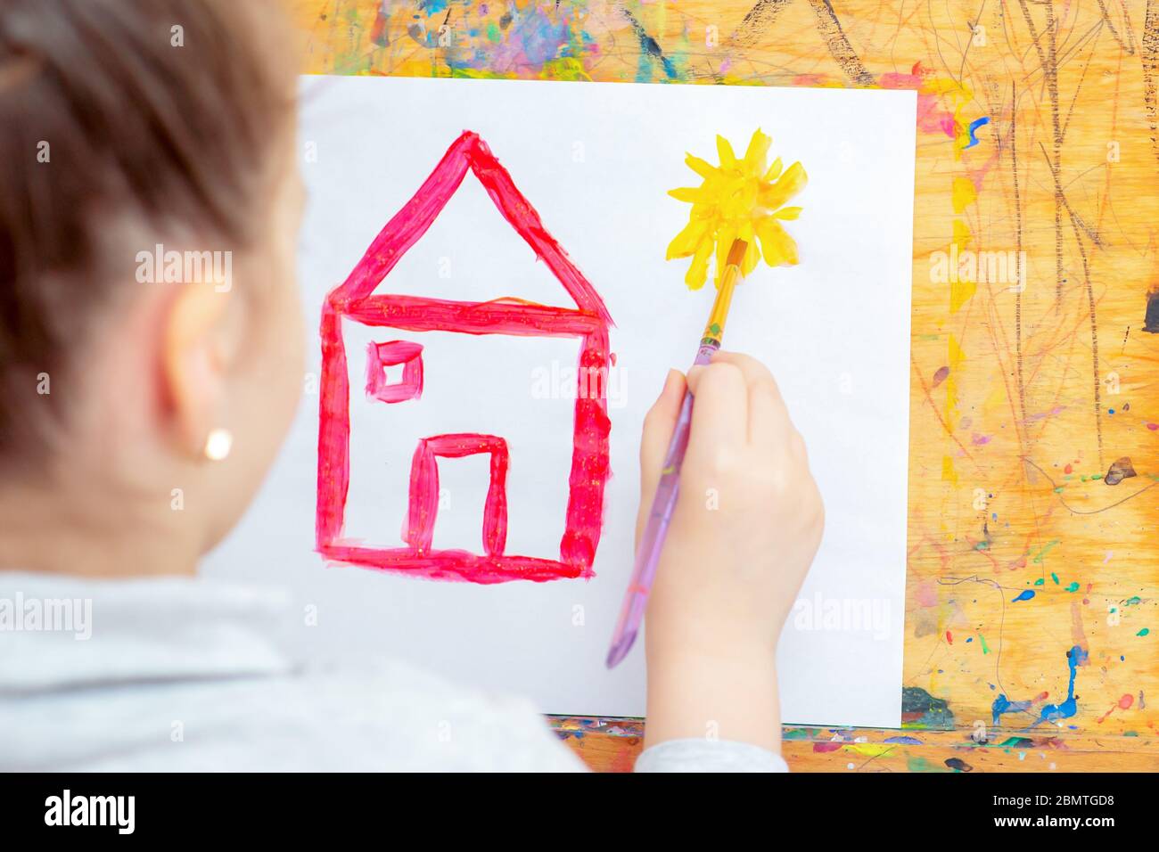 Hand of Little girl drawing yellow sun and red house with brush on easel at home. Children's creativity. Stock Photo