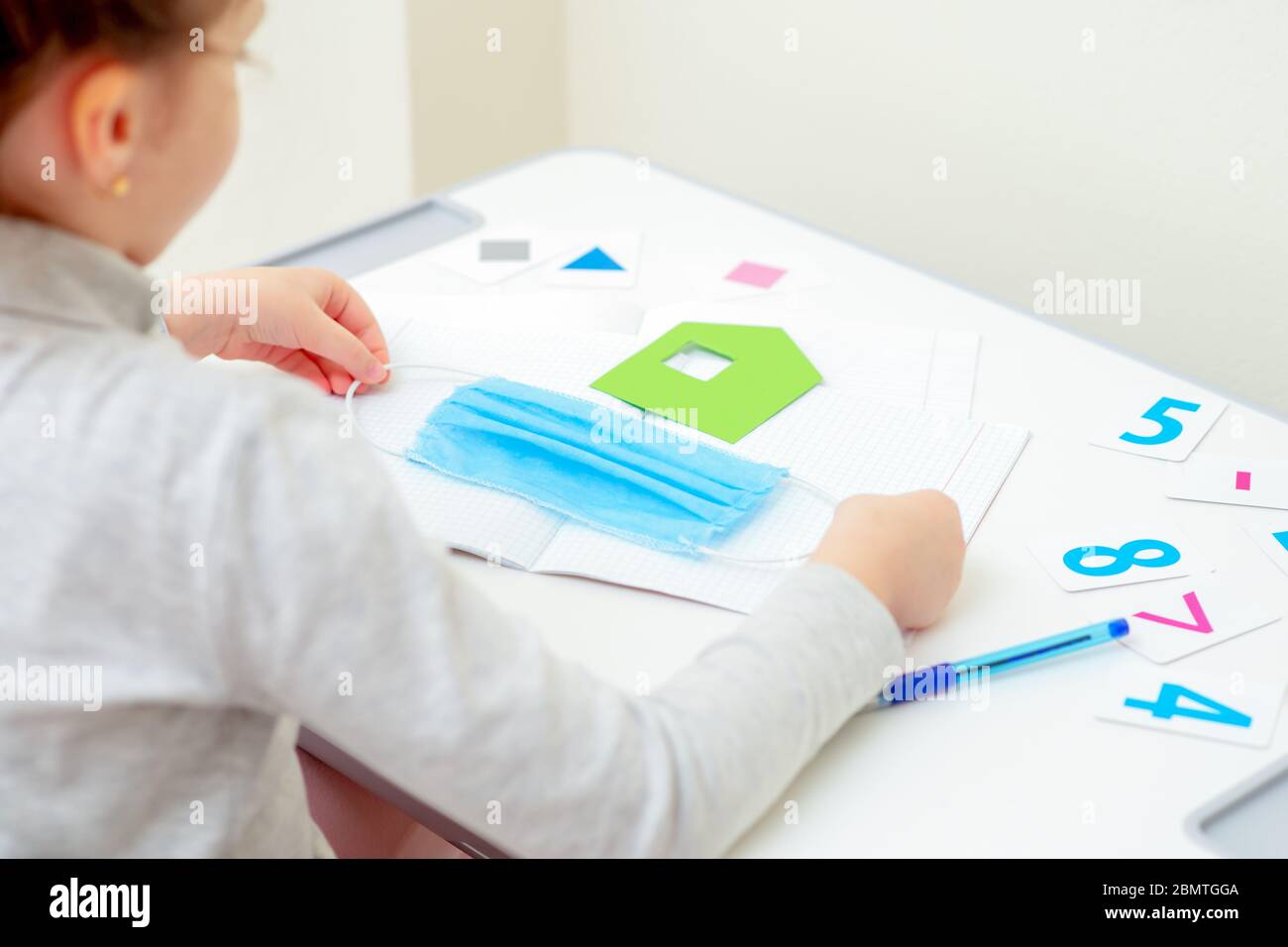 Little girl is holding medical mask for protection near paper green house over copybook on white desk at home. Quarantine studying concept. Stock Photo