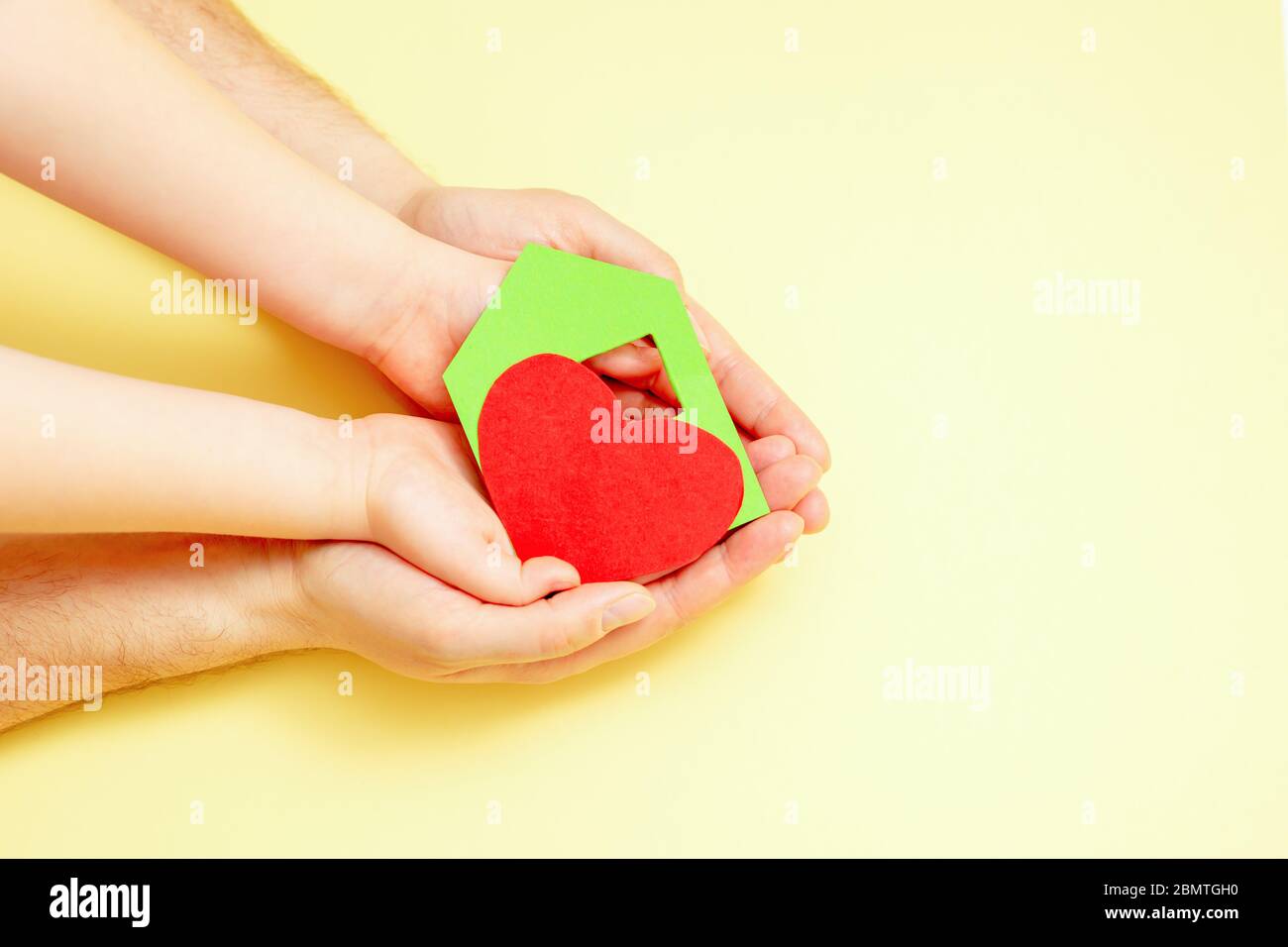 Paper green house with red heart in dad and child's hands on yellow background. Charity concept. Stock Photo