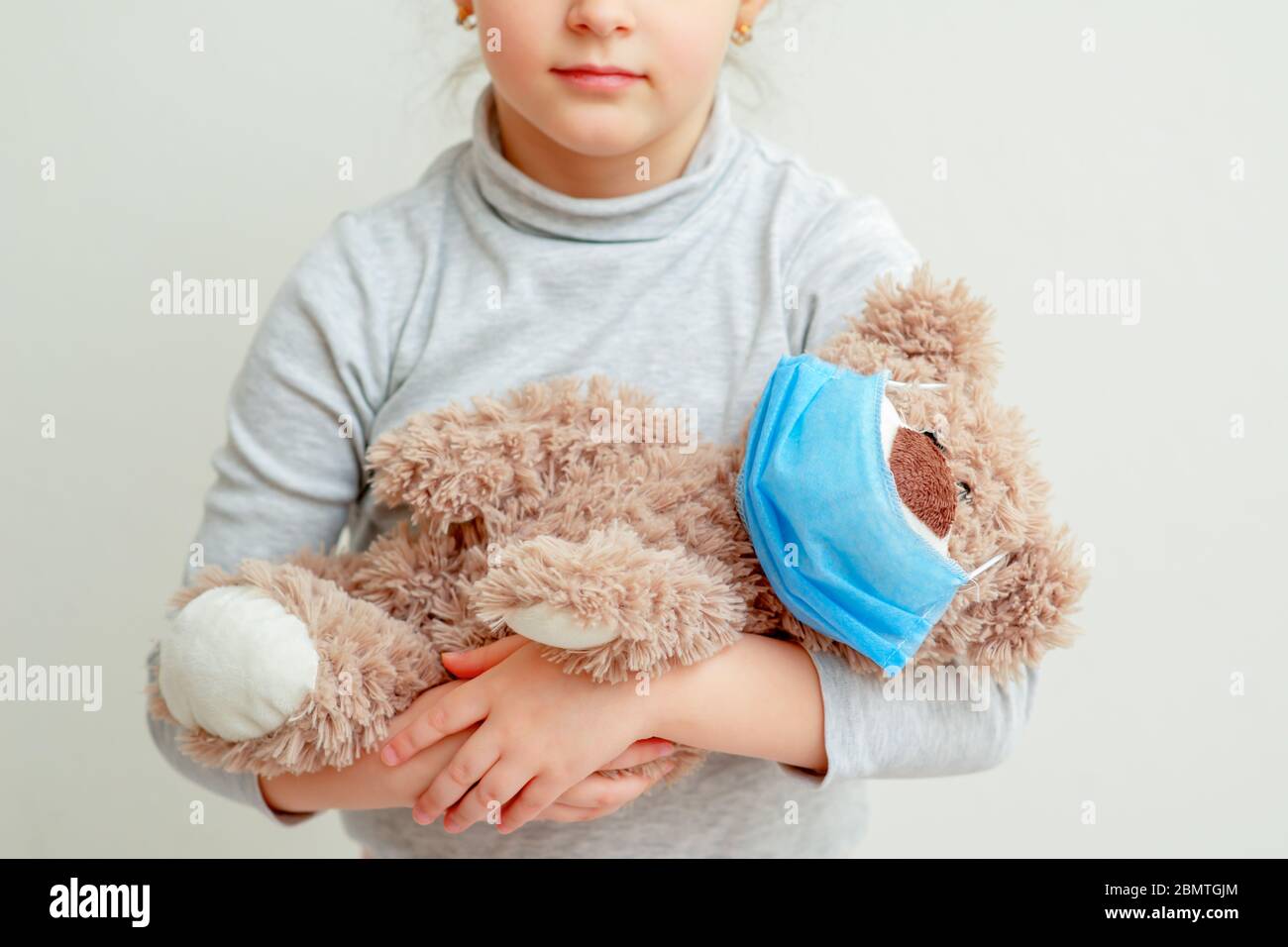 Toy bear with a medical mask in hands of sad little girl during quarantine on light background. Quarantine weekdays of children. Stock Photo