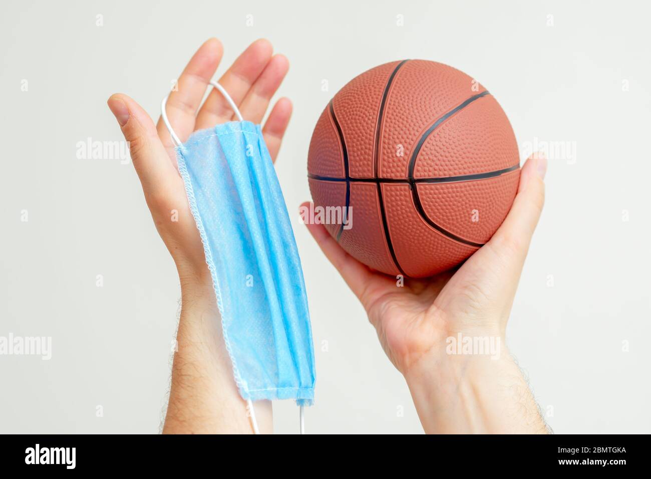 Closeup of basketball ball with a medical mask in man's hands on a light background. Cancellation of sporting events. Stock Photo