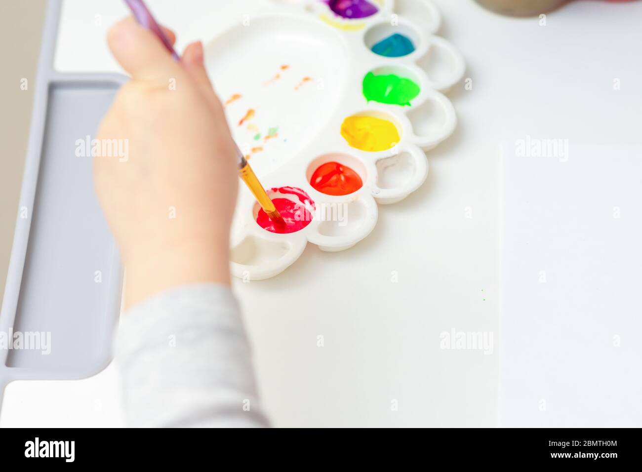The child is ready to paint with watercolors. Child's hand holding brush over palette on blank sheet of white paper with watercolors. Stock Photo