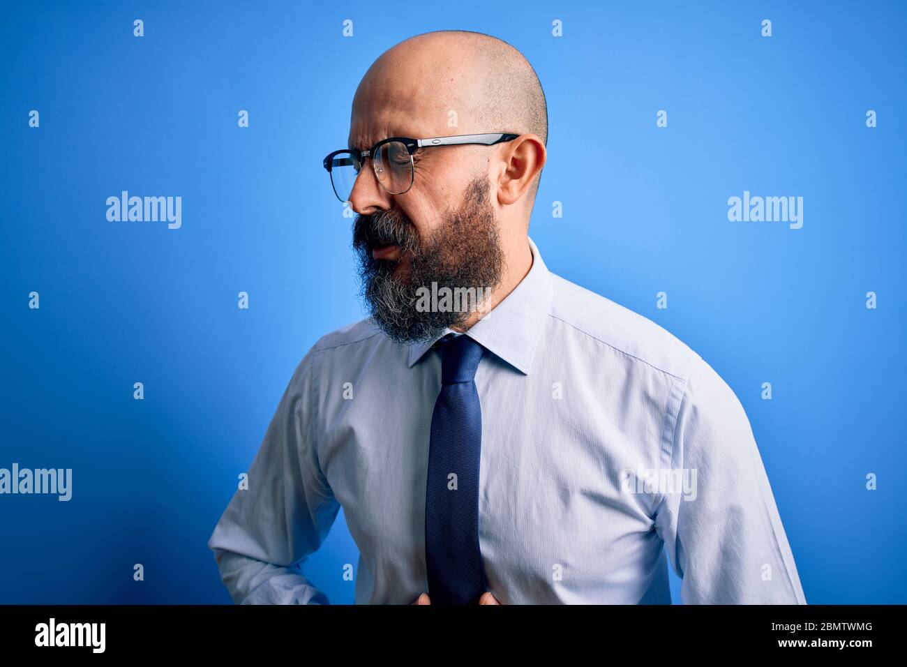 Handsome business bald man with beard wearing elegant tie and glasses over blue background with hand on stomach because nausea, painful disease feelin Stock Photo
