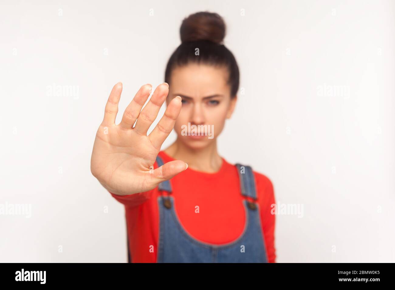Forbiddance, prohibition symbol. Portrait of strict girl with hair bun in denim overalls doing stop sign with raised hand, warning expression with bos Stock Photo