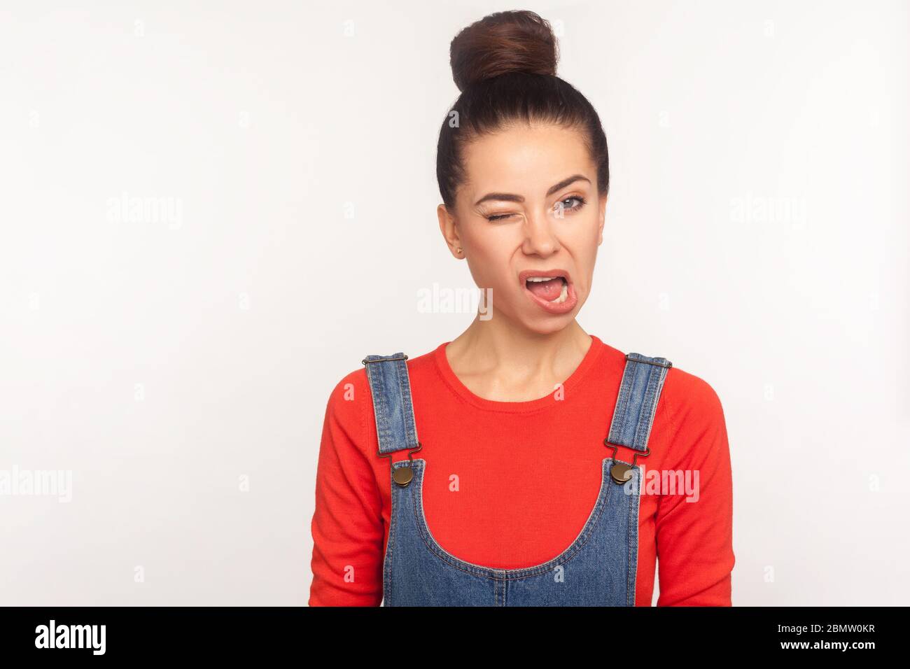 Portrait of funny displeased girl with hair bun in denim overalls winking to camera, looking with ugly skewed face, nasty dissatisfied grimace, expres Stock Photo