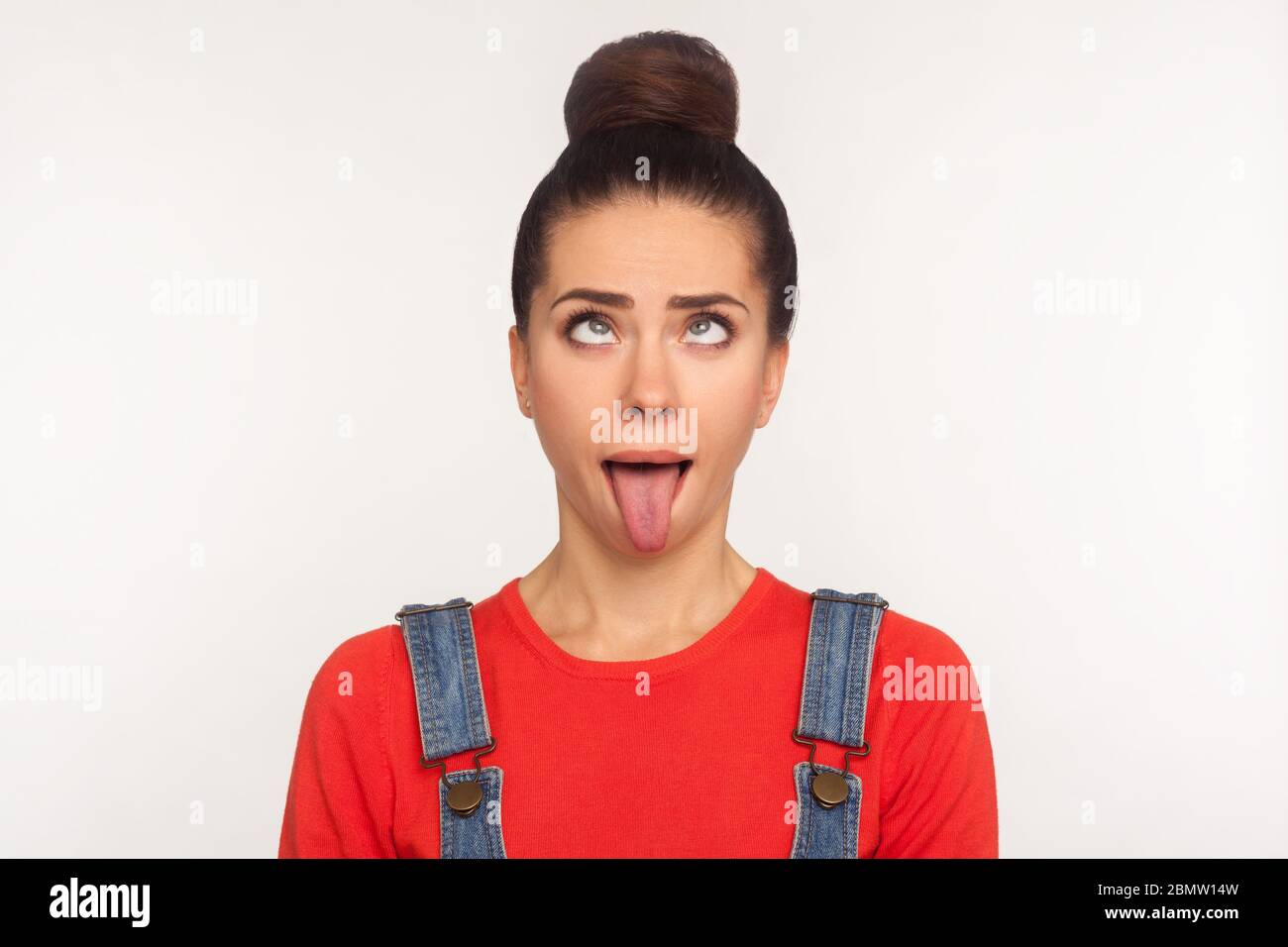 Closeup of amusing funny girl with hair bun looking up with eyes crossed and demonstrating tongue out, aping making dumb stupid face, awkward brainles Stock Photo