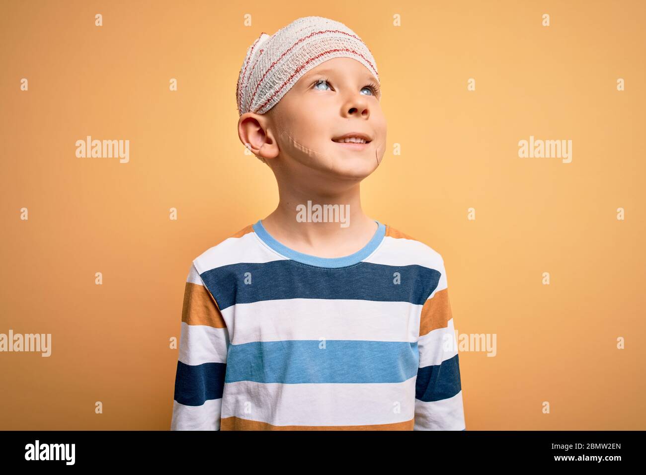 Young little caucasian kid injured wearing medical bandage on head over yellow background looking away to side with smile on face, natural expression. Stock Photo