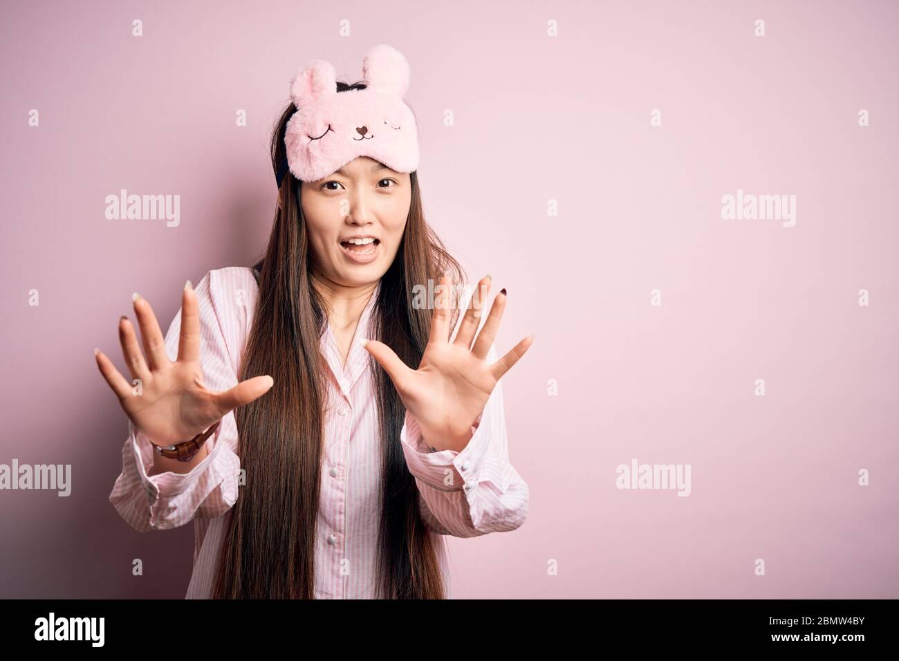 Young asian woman wearing pajama and sleep mask over pink isolated background afraid and