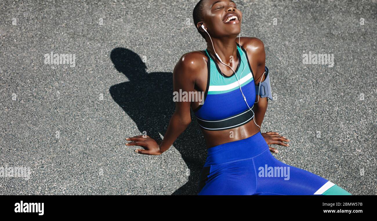 Top view shot of happy woman sitting on road after a morning run. Fit woman on road taking a break after outdoor workout. Stock Photo