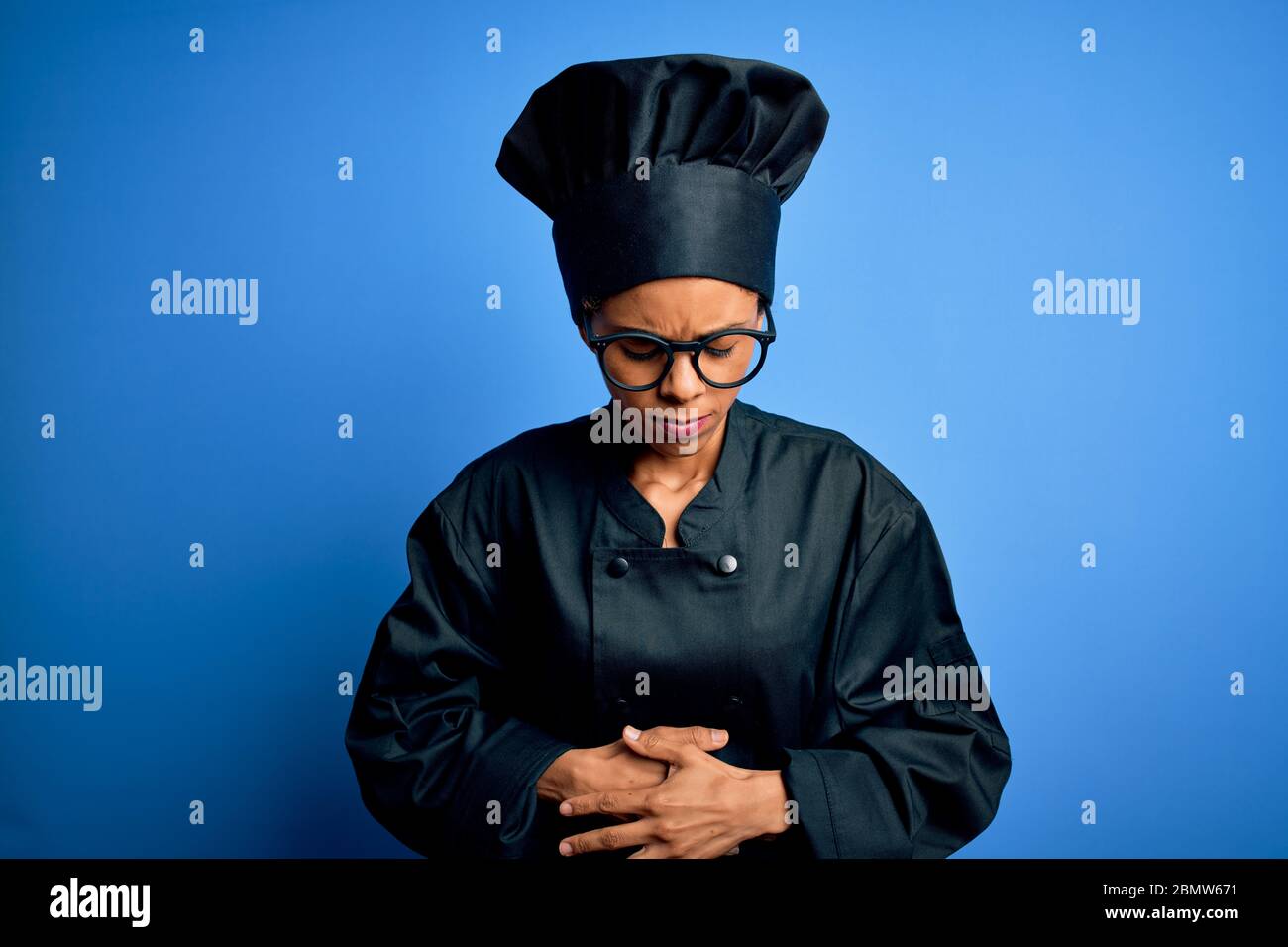 Young african american chef woman wearing cooker uniform and hat over blue background with hand on stomach because indigestion, painful illness feelin Stock Photo