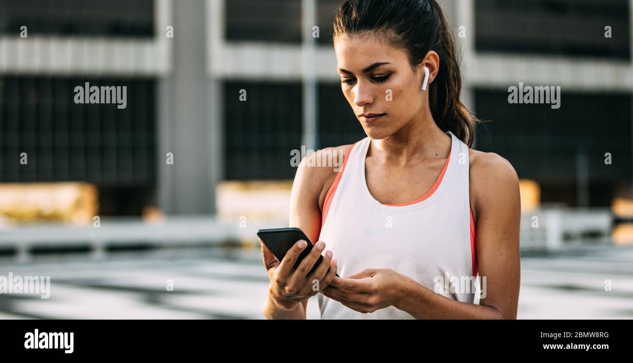 Female in sportswear using her smart phone after workout session. Healthy woman wearing earphones taking a break from workout and checking her fitness Stock Photo