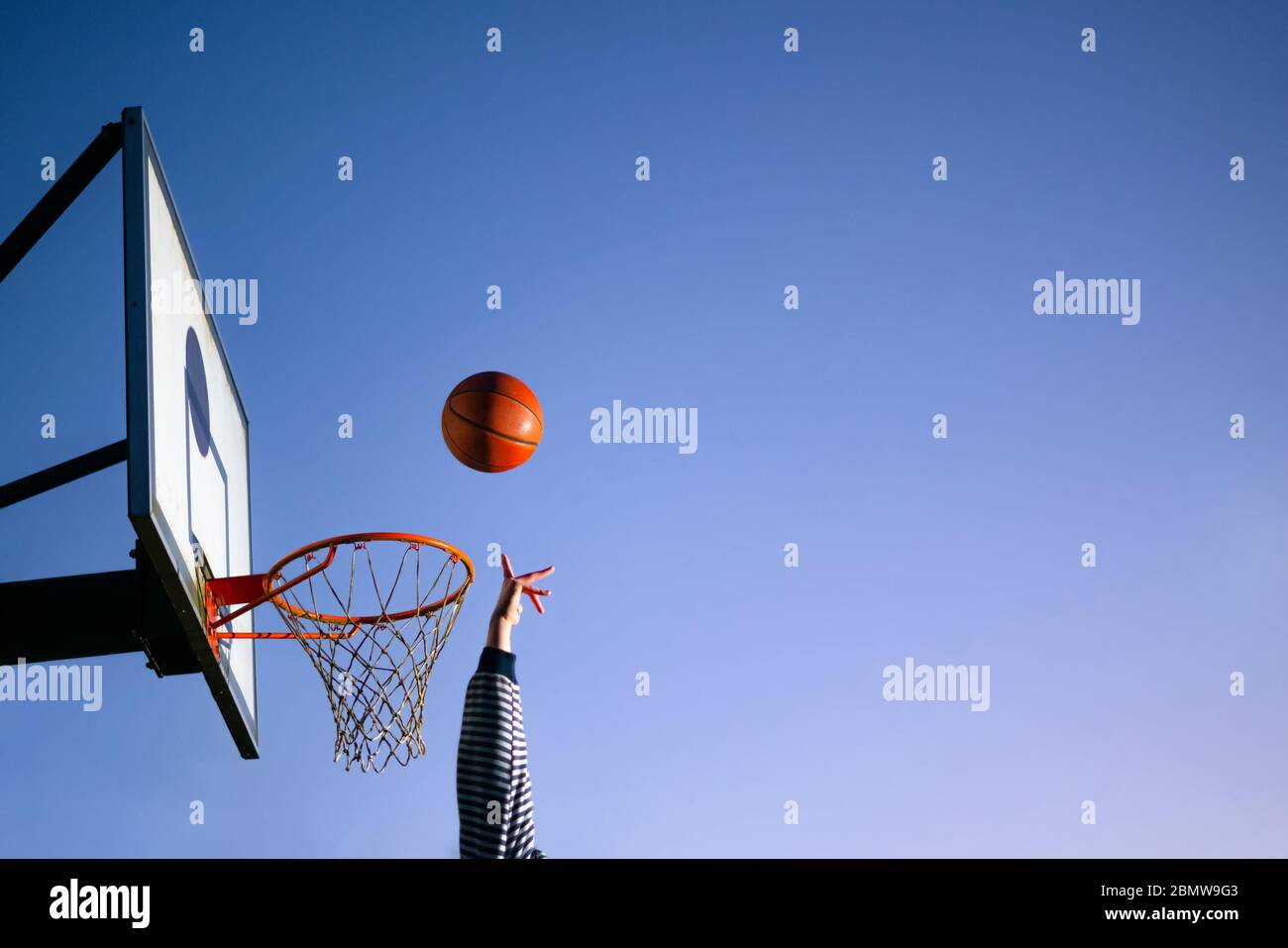 Street basketball ball player throwing ball into the hoop. Close up of hand, orange ball above the hoop net with blue sky in the background. Concept o Stock Photo