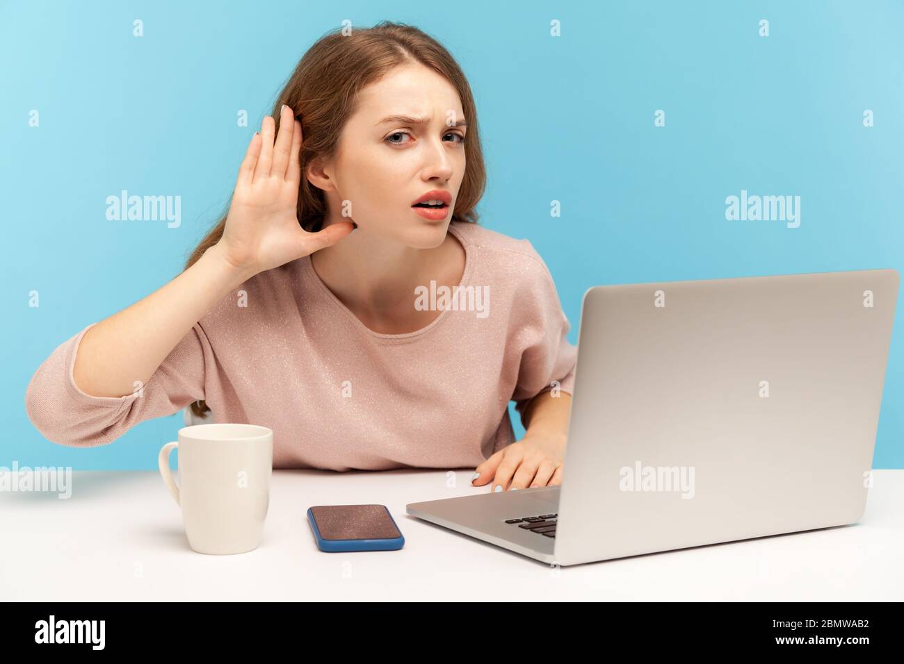 What? I can't hear you! Female office worker sitting at desk with laptop and keeping hand at her head, listening carefully intently to secret gossip. Stock Photo