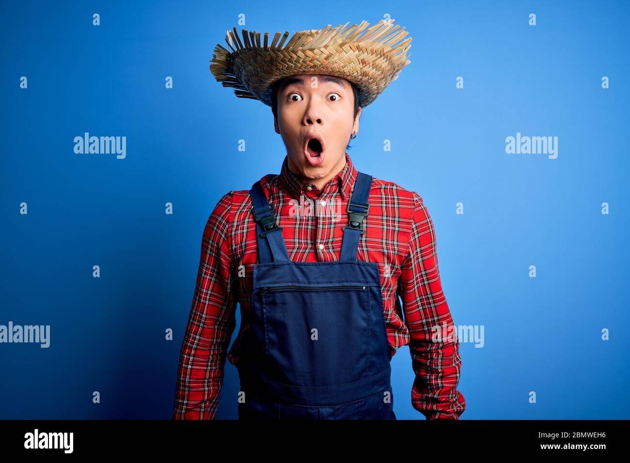 Young handsome chinese farmer man wearing apron and straw hat over blue ...