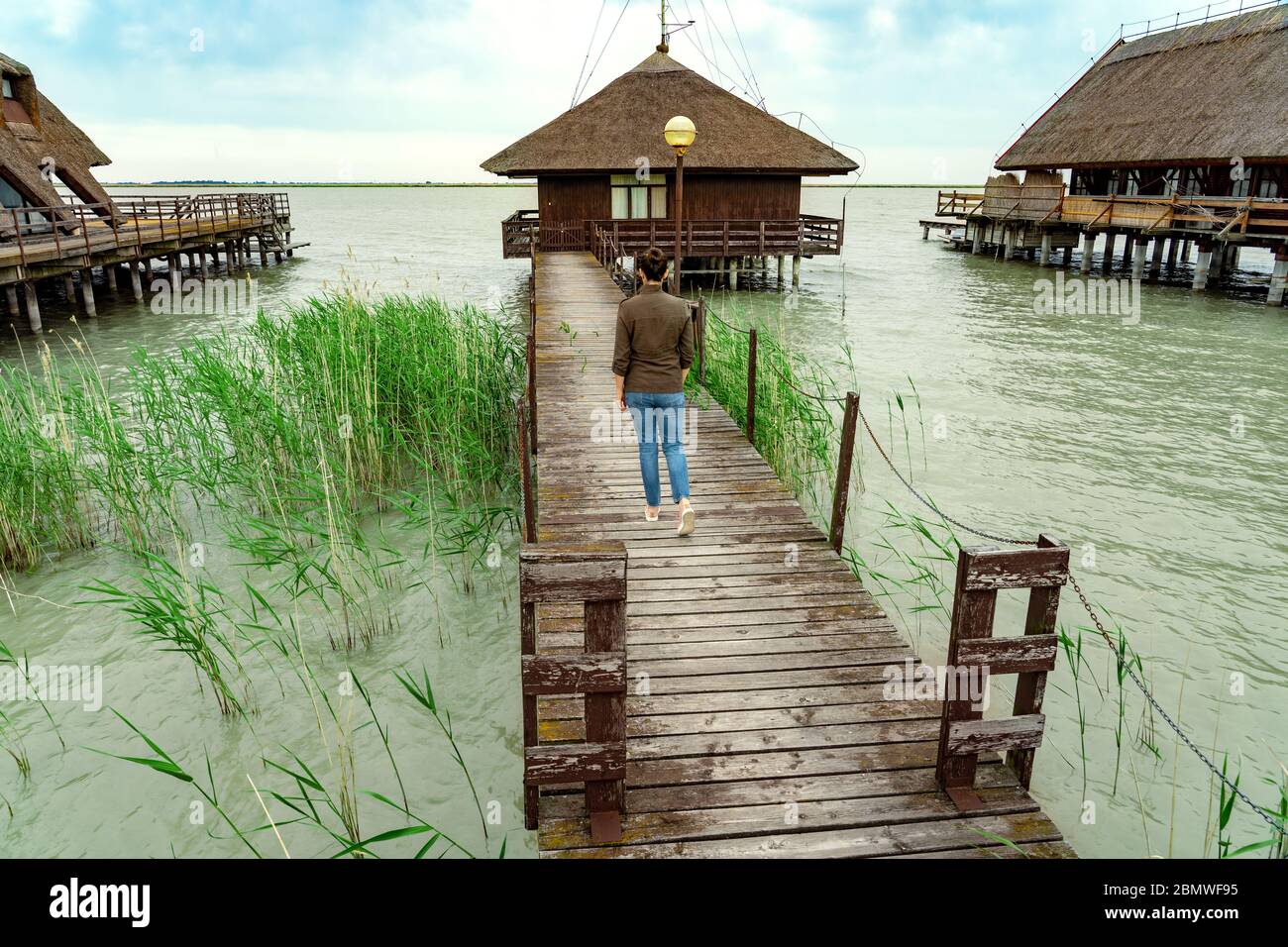 peaceful nature at the Lake Fertő in Hungary with wooden pier bungalows cabins on the lake and straw in the water with a tourist woman walking on the Stock Photo