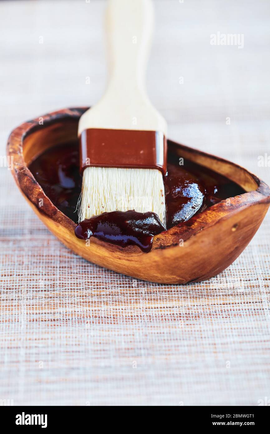 BBQ sauce on the tip of a basting brush with a wooden bowl filled with the condiment. Selective focus with blurred background. Stock Photo