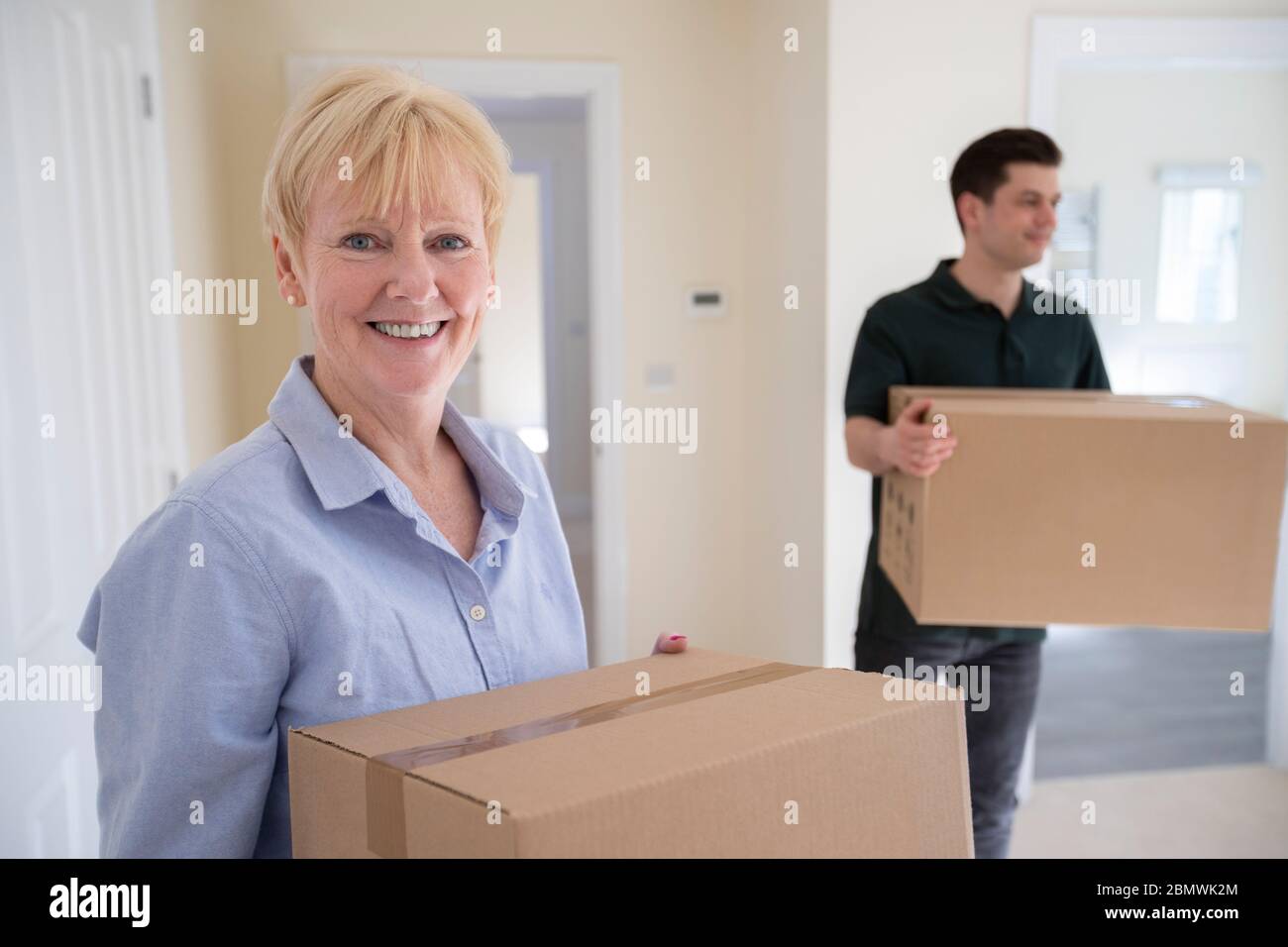 Portrait Of Senior Woman Downsizing In Retirement Carrying Boxes Into New Home On Moving Day With Removal Man Helping Stock Photo