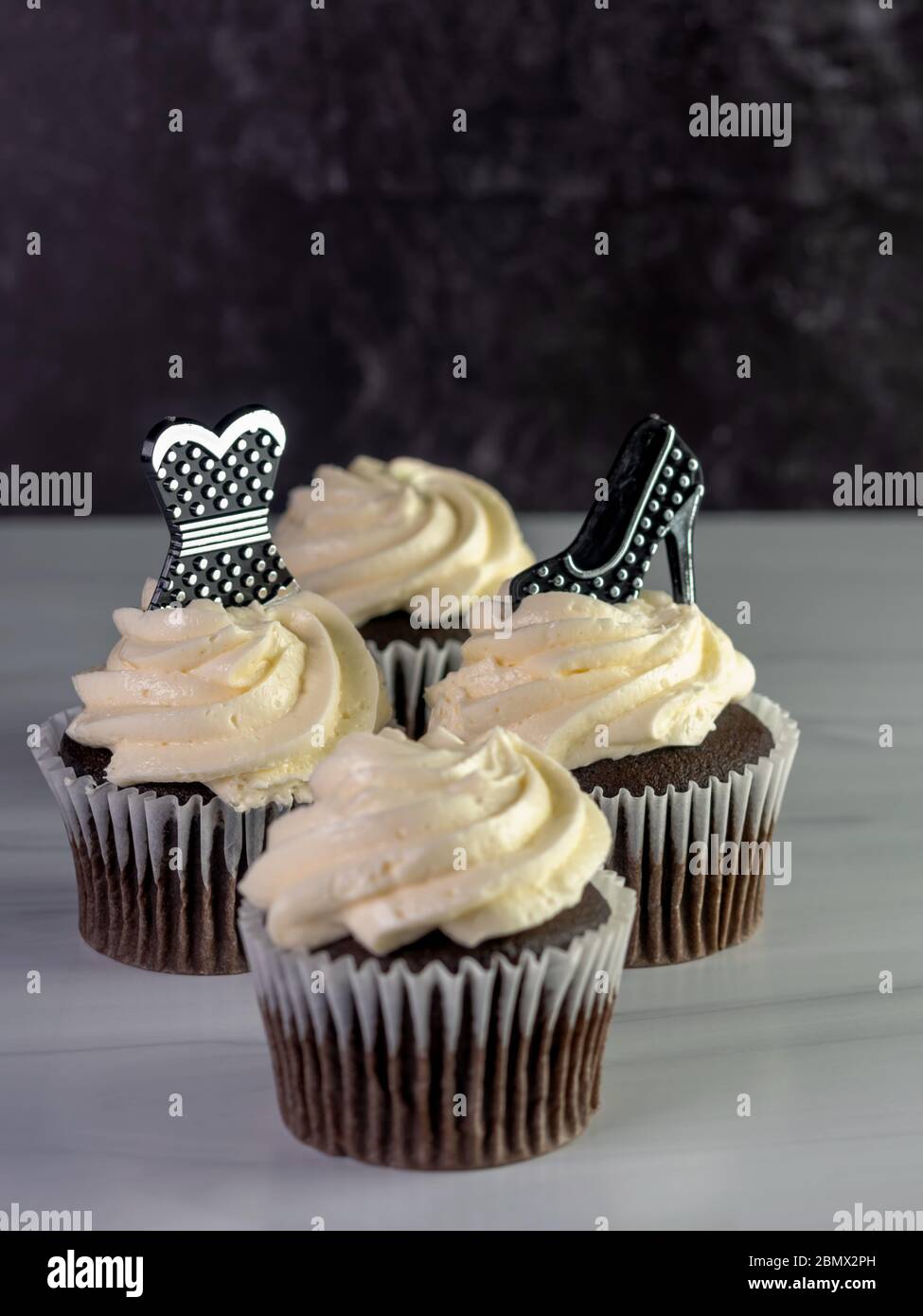 4 chocolate cupcakes with white frosting swirled high with a black and silver polka dot high heel shoe and prom dress garnishing them, on a white coun Stock Photo