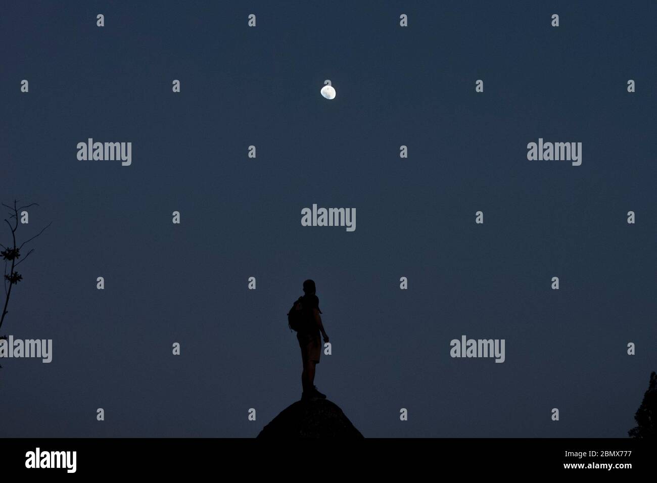 Man stands backlit on a rock looking up to an almost full moon, with the blue night sky as a background Stock Photo