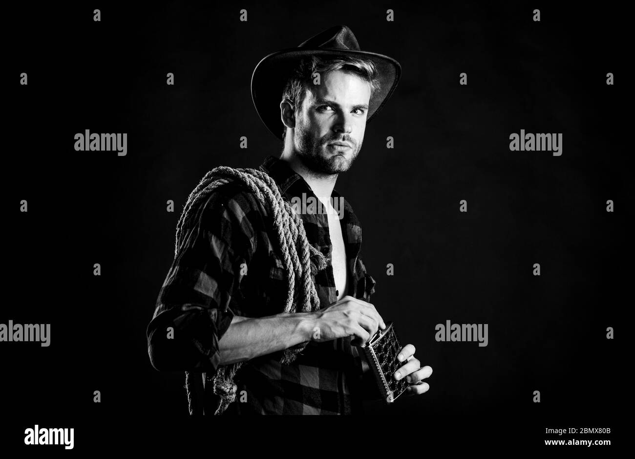 Western culture. Man wearing hat hold rope and flask. Lasso tool of American cowboy. Sheriff concept. Brutal cowboy drinking alcohol. Man handsome unshaven cowboy black background. Western life. Stock Photo