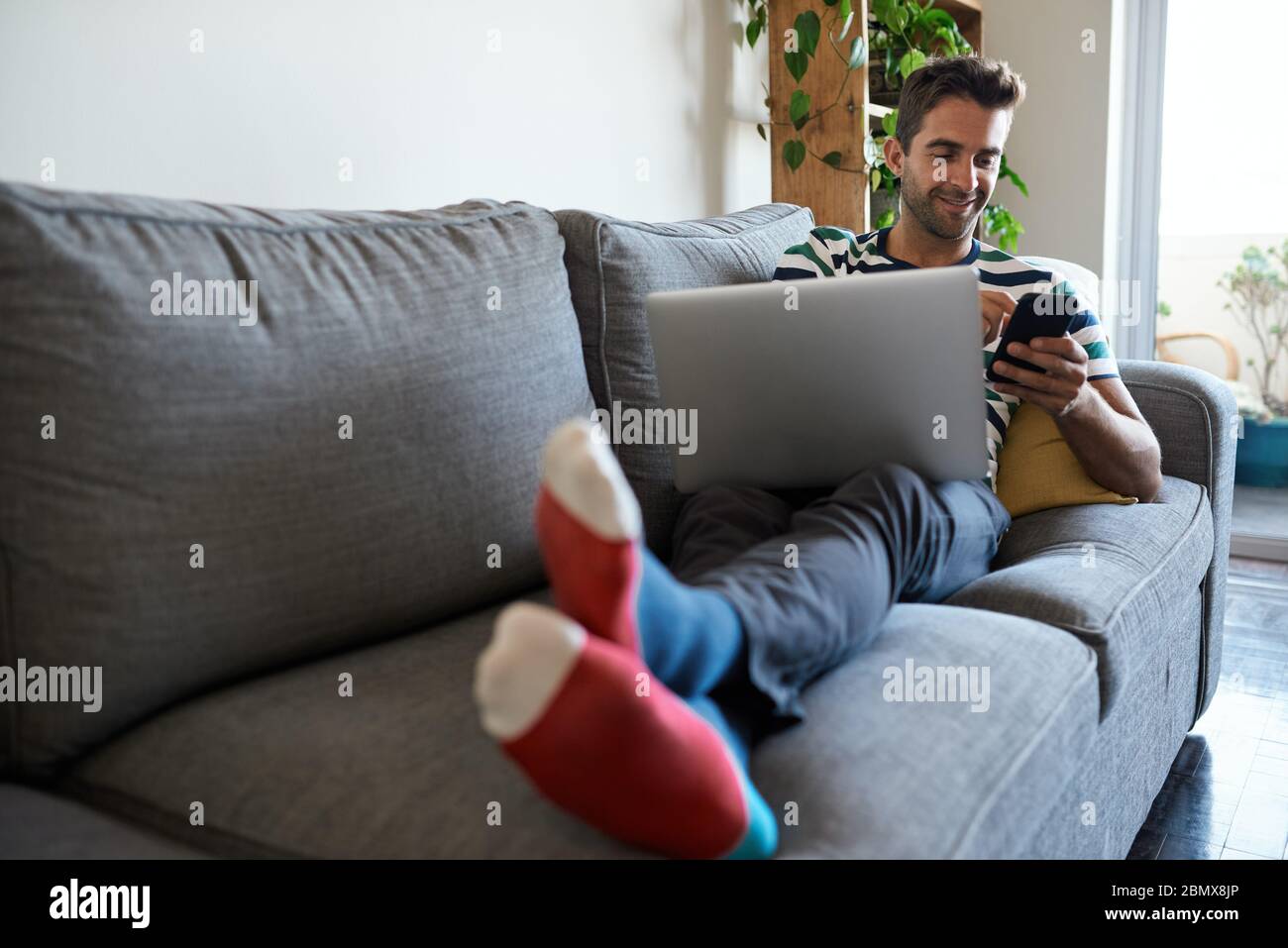 Smiling man using a cellphone and laptop on his sofa Stock Photo