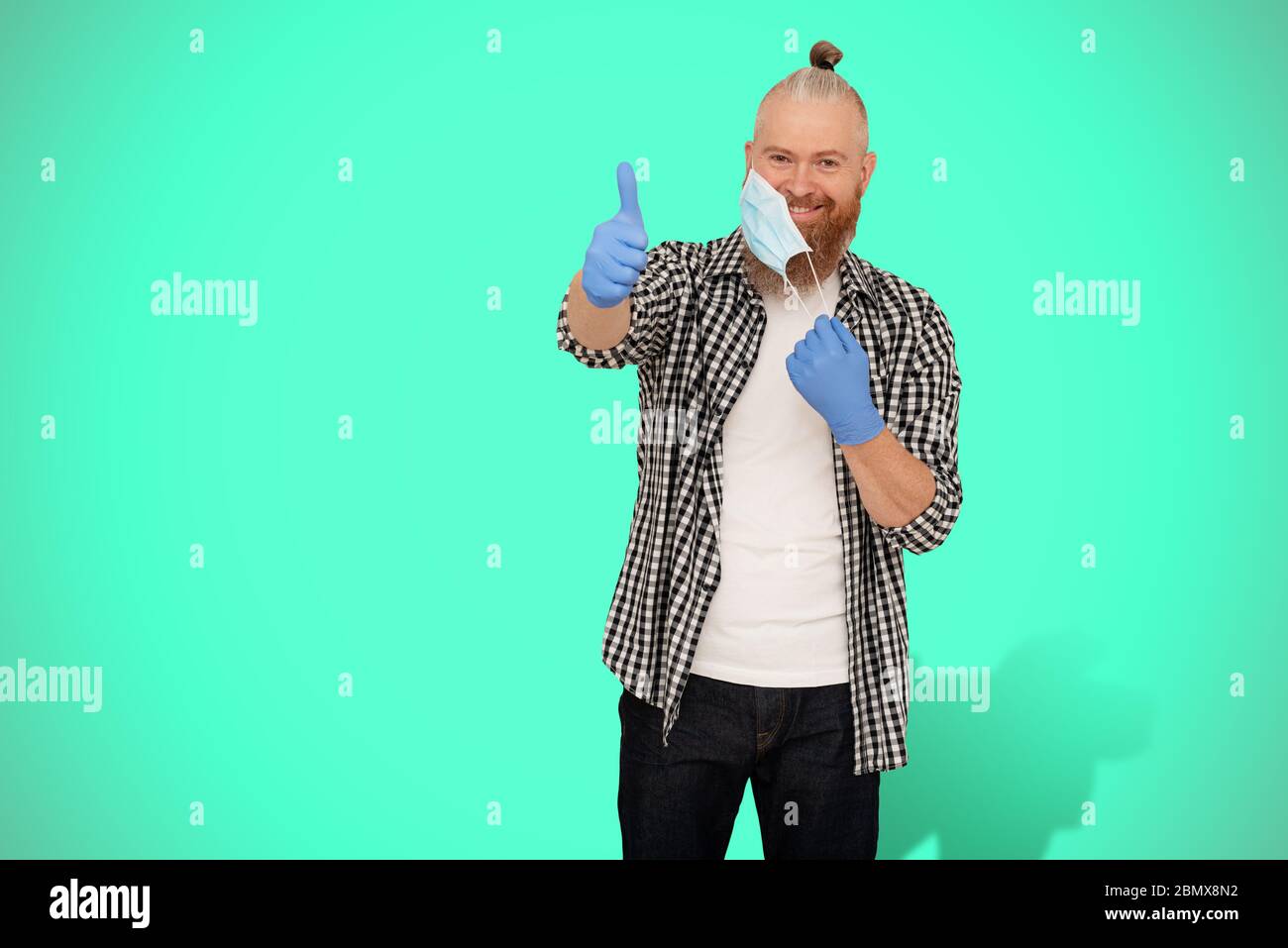 Happy positive smiling bearded man taking off protective medical mask and showing thumbs up on neo mint background. Happy end of quarantine Stock Photo