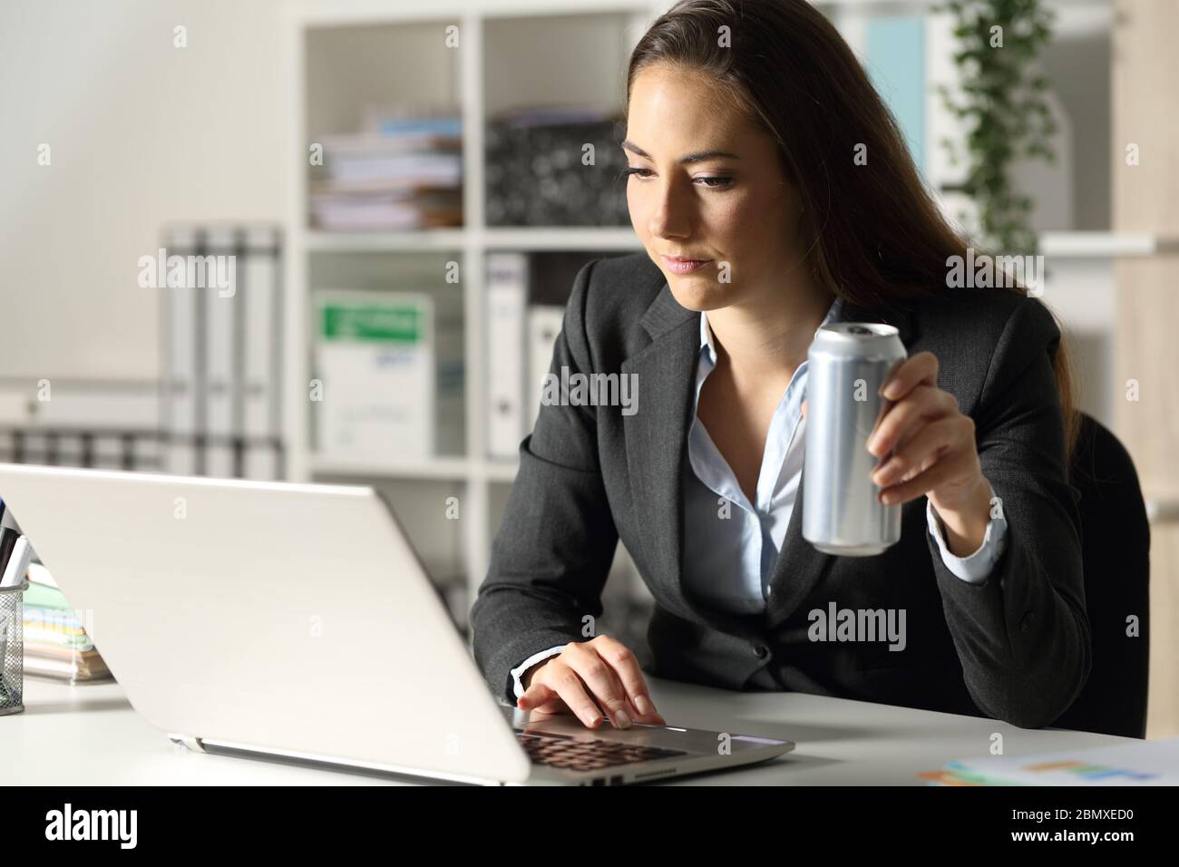 Executive woman holds energy drink can working late hours on laptop at night in the office Stock Photo