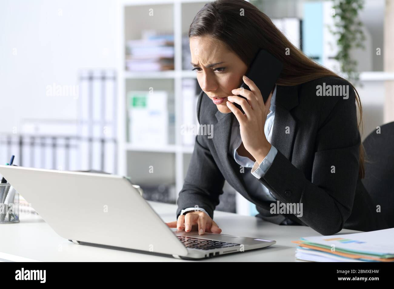 Worried executive woman calling on smart phone checking laptop sitting on her desk at office Stock Photo