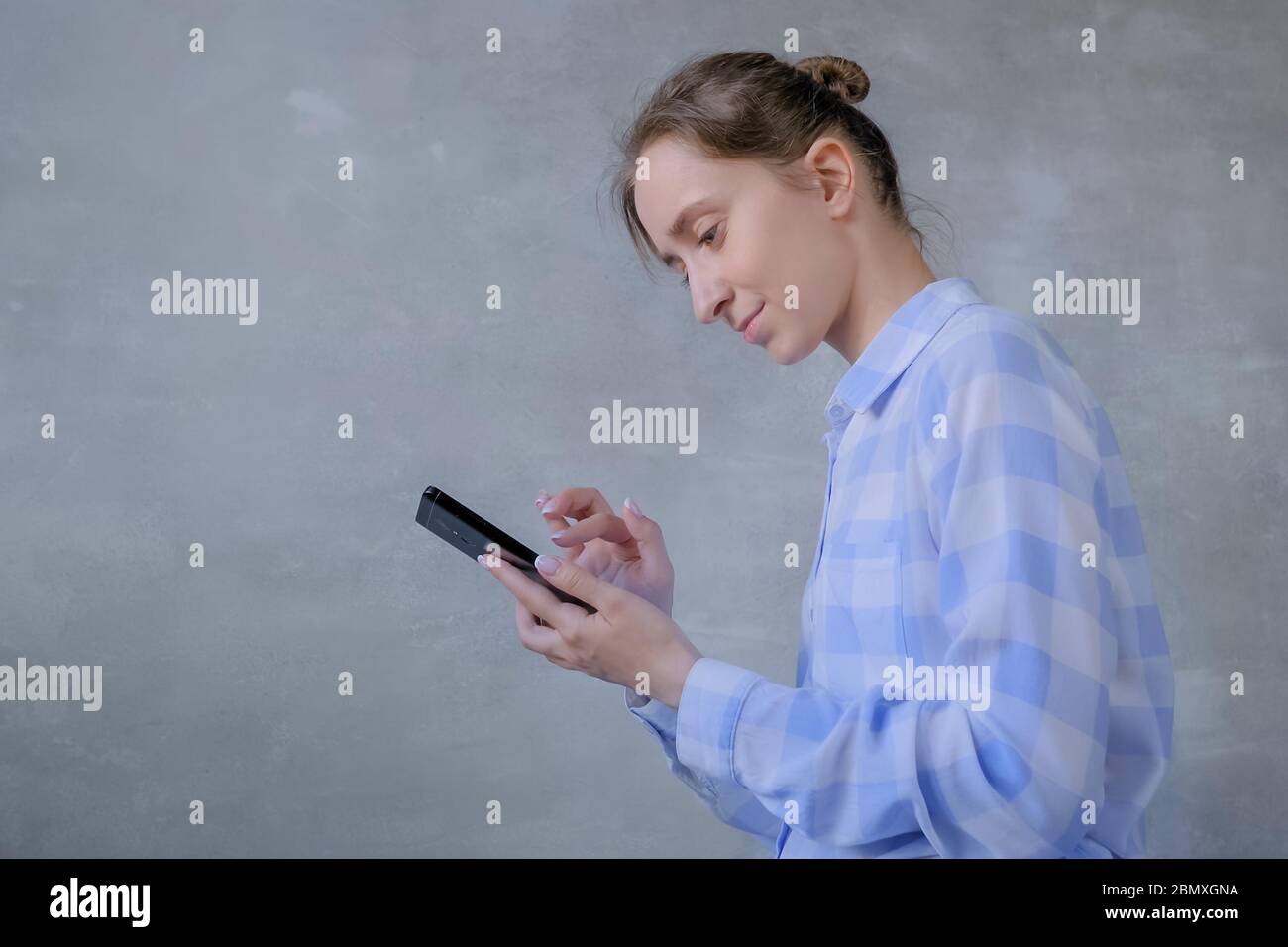 Portrait of woman in blue and white plaid shirt using black smartphone with touchscreen display in room with grey wall - scrolling and touching Stock Photo