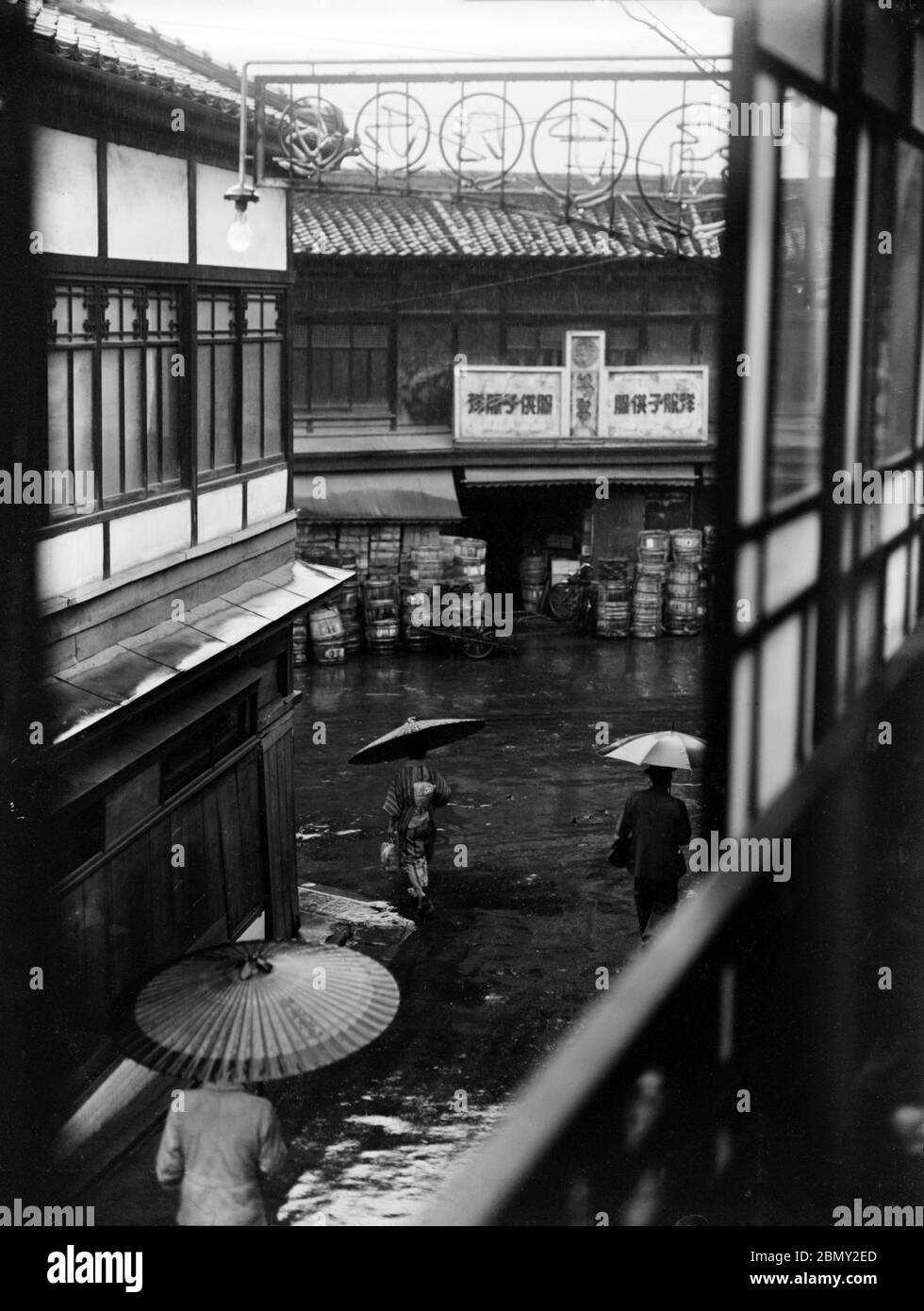 [ 1930s Japan - Backstreet View ] —   View from a second storey backstreet room during a rainy day somewhere in Japan. Two people with Japanese paper parasols and one person with a Western umbrella are walking down the street. In the back a shop can be seen.  20th century vintage glass negative. Stock Photo