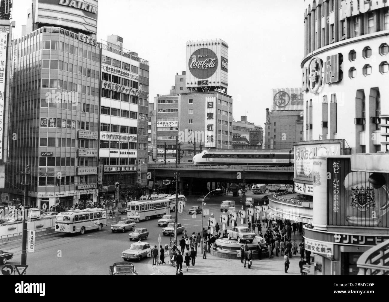 [ 1960s Japan - Yuraku-cho, Tokyo ] —   Taxis, buses and streetcars pass in front of the Nihon Gekijo (日本劇場) in Yuraku-cho (有楽町) in Chiyoda-ku, Tokyo in 1967 (Showa 42).  In the back, a new 0 series Shinkansen 'Bullet Train' can be seen.  The Tokaido Shinkansen began service on October 1, 1964 (Showa 39), just in time for the first Tokyo Olympics.  20th century gelatin silver print. Stock Photo