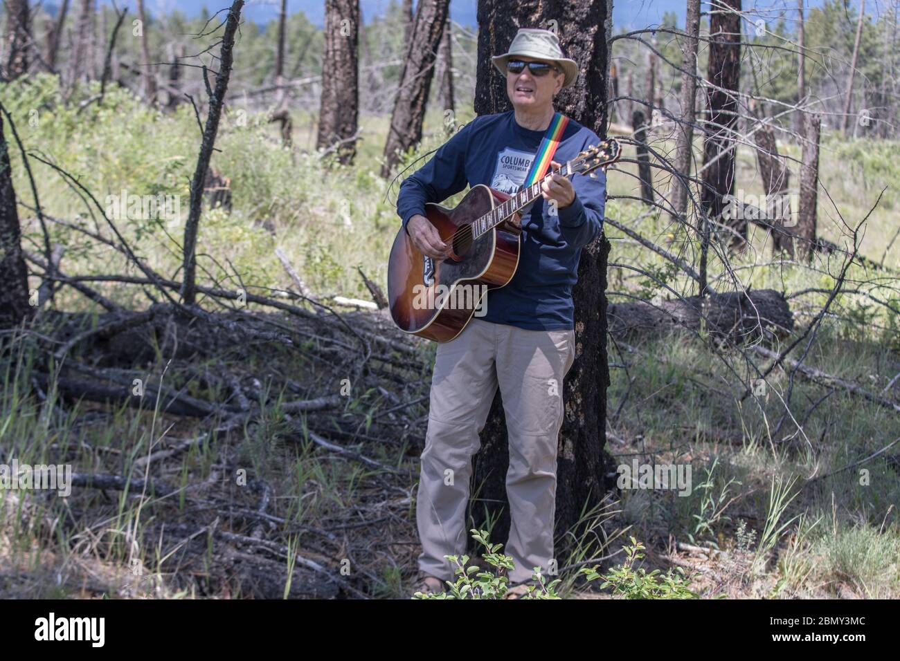Man playing guitar,  outdoors, standing in grass field, with blue sky and clouds. Stock Photo