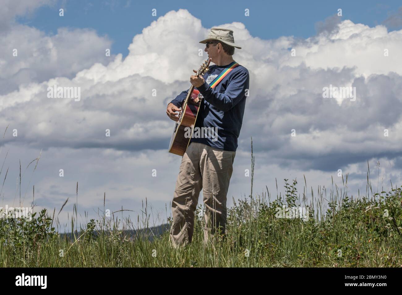 Man playing guitar,  outdoors, standing in grass field, with blue sky and clouds. Stock Photo