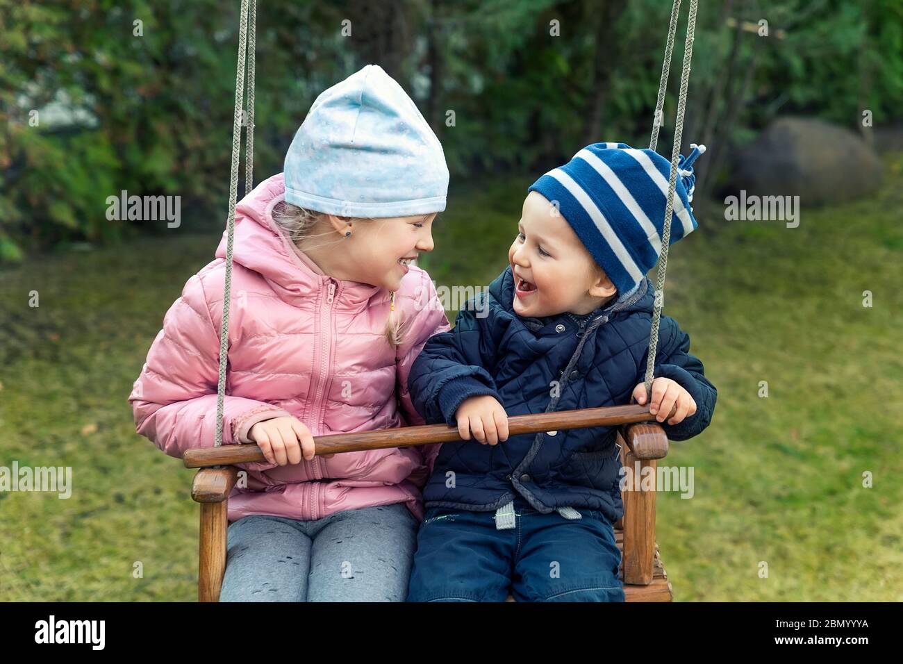 Two cute adorable playful caucasian siblings boy girl child enjoy having fun swinging wooden swing at backyard together with father. Little toddler Stock Photo