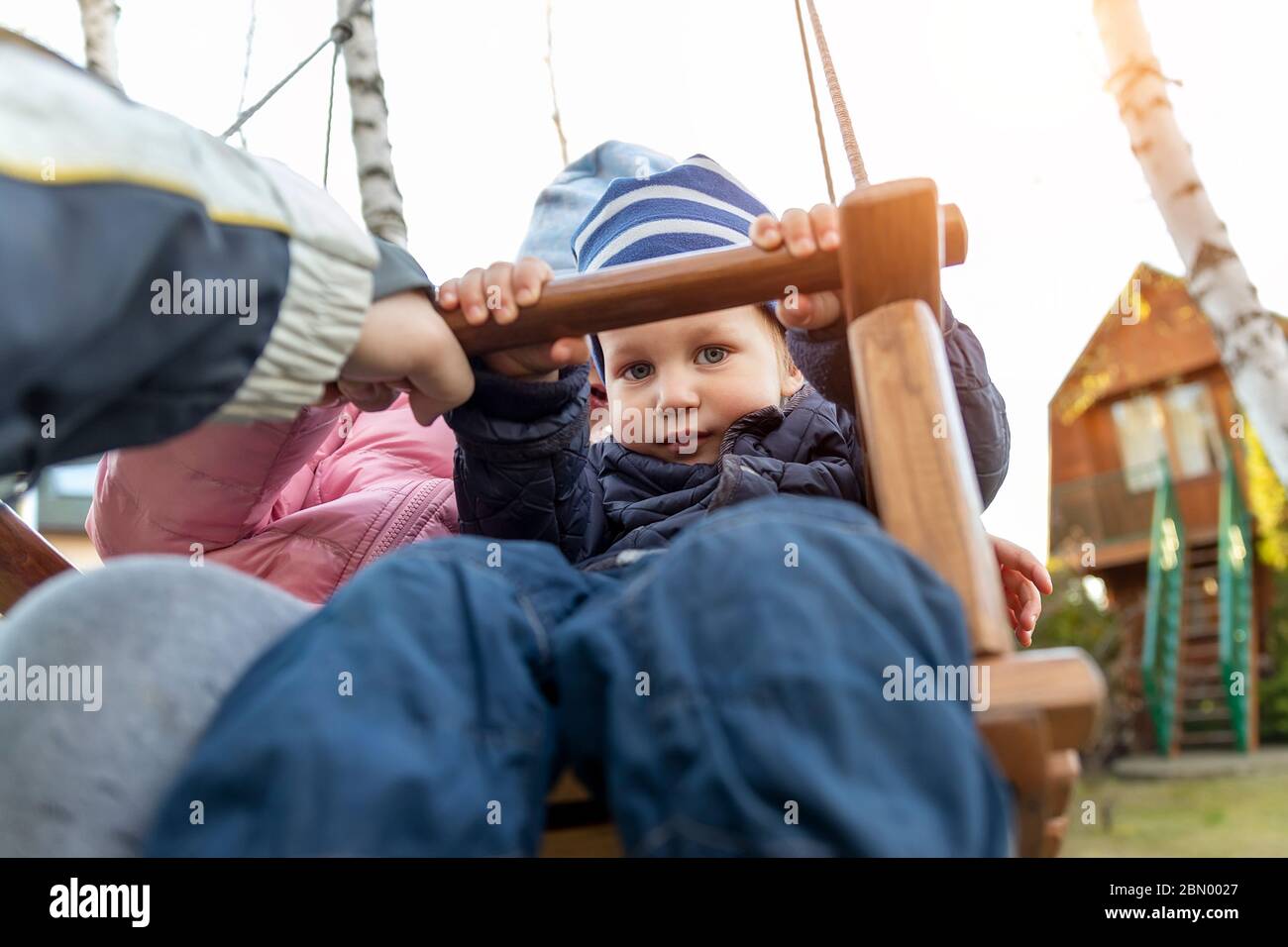 Two cute adorable playful caucasian siblings boy girl child enjoy having fun swinging wooden swing at backyard together with father. Little toddler Stock Photo