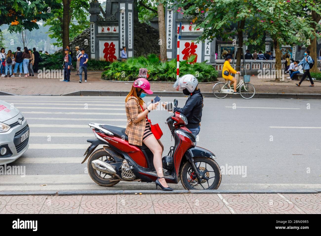 A young woman sitting on a motorbike and a young man standing each wearing face masks and helmets look at their mobile phones in Hanoi, north Vietnam Stock Photo