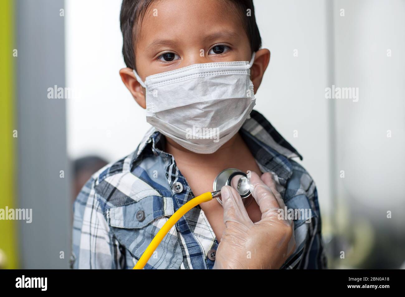 Child wearing a medical mask to prevent spread of virus is getting a heart screening with stethoscope placed on chest. Stock Photo