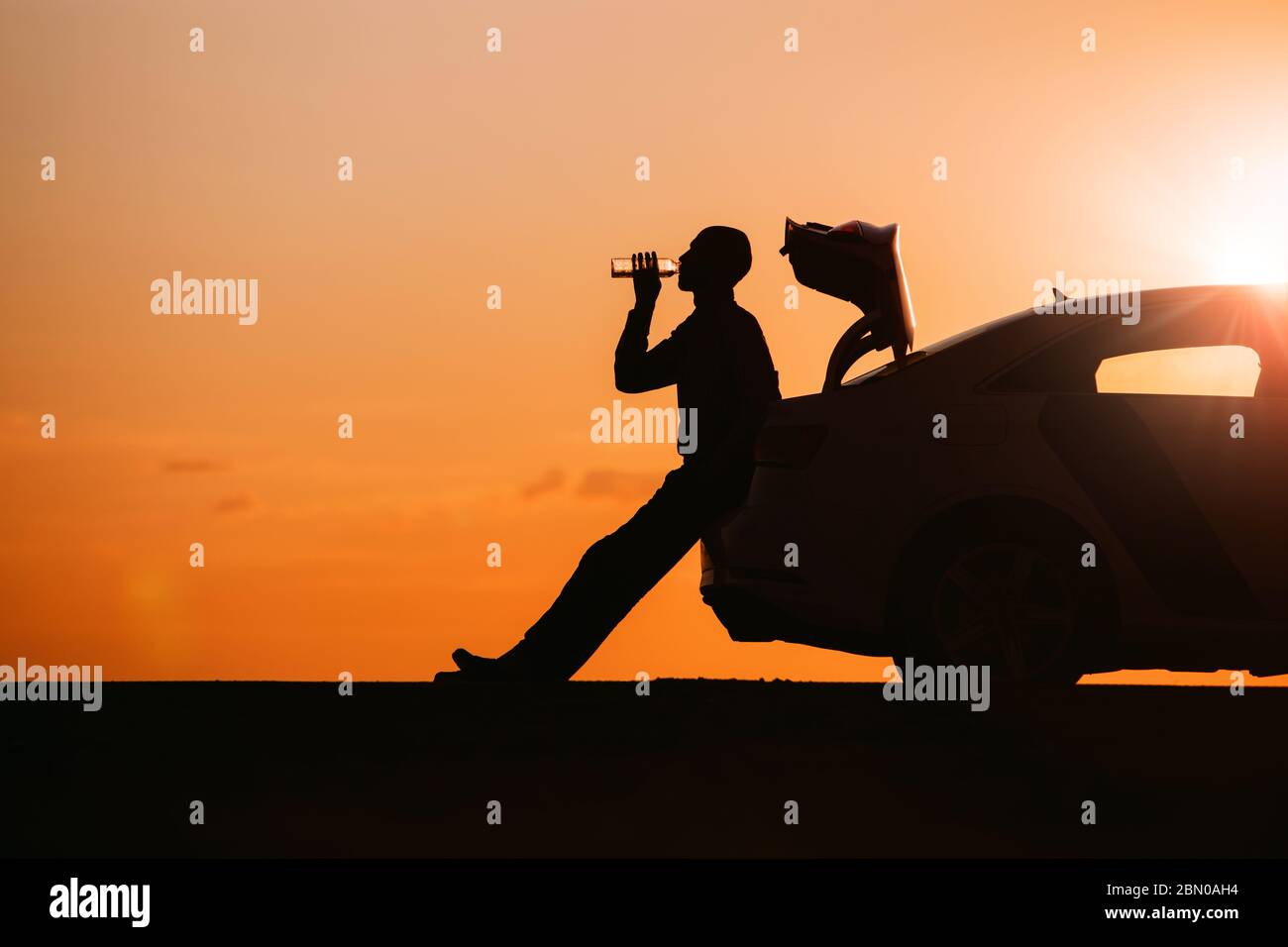 Silhouette of man driver relaxing after a ride, sitting on the trunk of his car and drinking water from a bottle, side view. Sunset time. Stock Photo