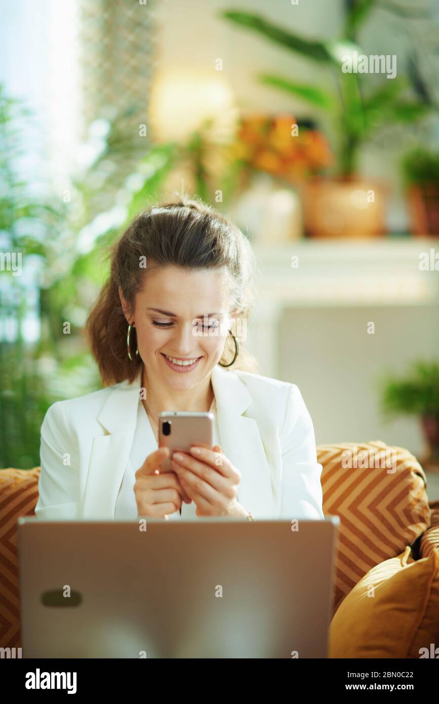 happy modern woman in white blouse and jacket with laptop and smartphone texting in the modern living room in sunny day. Stock Photo