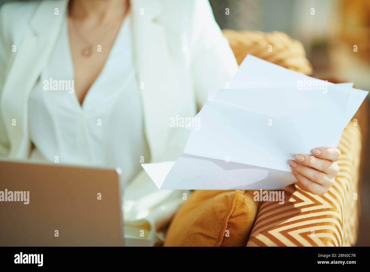 Closeup on woman with documents using laptop while sitting on divan in the house in sunny day. Stock Photo