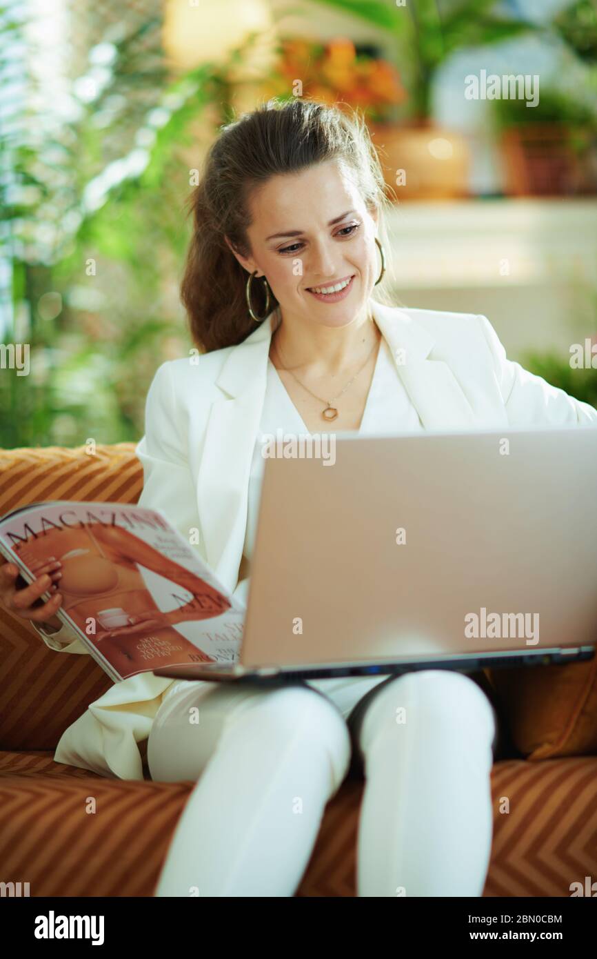 smiling modern middle age woman in white blouse and jacket with laptop and magazine at modern home in sunny day. Stock Photo