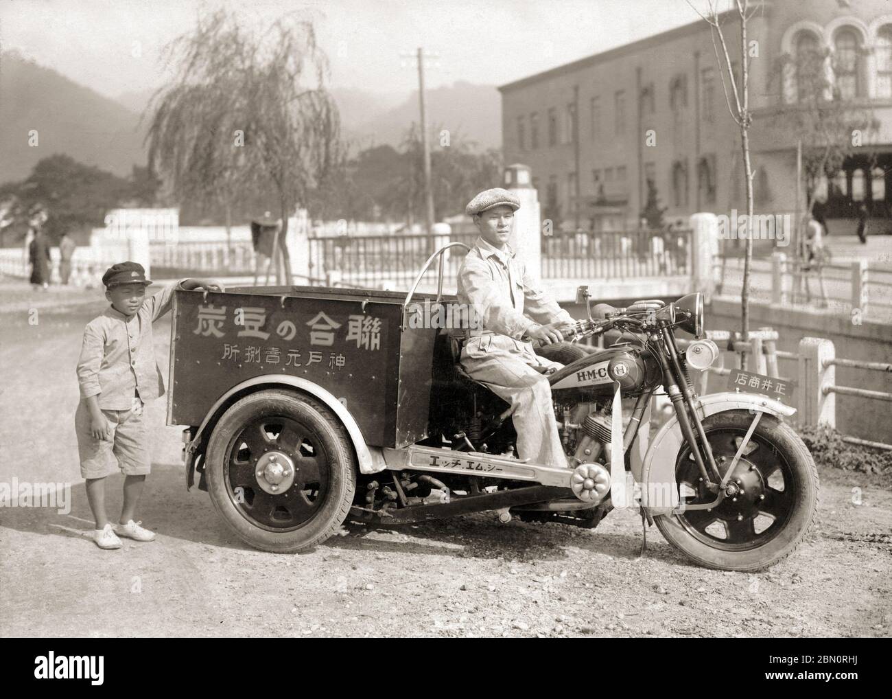 [ 1930 Japan - HMC Rear Car Motorcycle, Kobe ] —   A man selling charcoal briquettes poses on a rear car delivery motorcycle in Kobe.   The motorcycle is a trike manufactured by the Hyogo Motors Company (HMC, 兵庫モータース). No driver's license was needed for trikes below 750cc, making them extremely popular in Japan.  The photo is dated May 12, 1930.  Very few Japanese cargo trikes survived the war, making this photo an important historical document.  20th century gelatin silver print. Stock Photo
