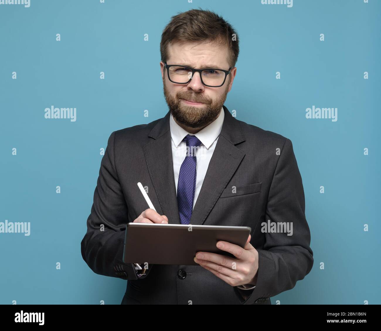 Bewildered businessman in a suit works with a tablet and stylus and looks thoughtfully at the camera with squinted eyes. Stock Photo